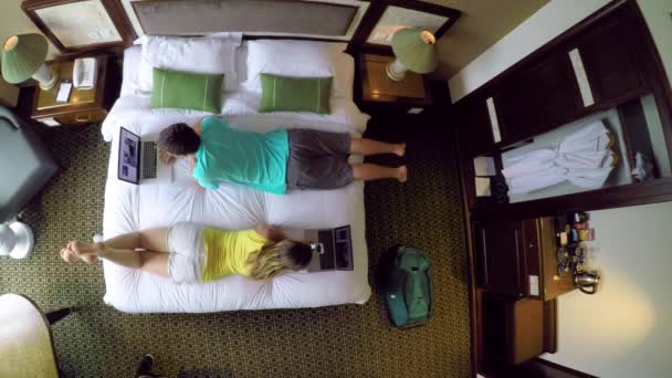 自上而下 男人在他的笔记本电脑上工作 而女朋友在越南度假期间摆弄她的智能手机 年轻的旅游夫妇使用他们的笔记本电脑 而躺在大舒适的床上 — 图库视频影像