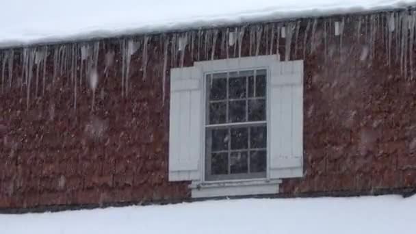 クローズアップ 雪のカリフォルニアの田舎の家の溝にぶら下がっている長いつらら 美しい白い木製の窓と牧歌的な建物を罰する吹雪 危険な雪が屋根と鋭い氷に覆われた — ストック動画