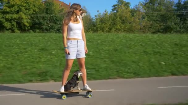 Vrolijke jonge vrouw rijdt een elektrische longboard met haar miniatuur pinscher hond — Stockvideo