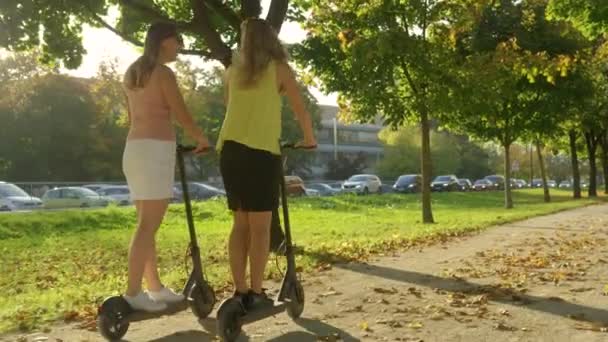LENS FLARE: Unga kvinnor skrattar när de rider elektriska skotrar längs natursköna avenyn — Stockvideo