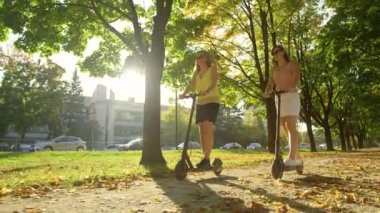 İki bayan arkadaş gülüyor ve yeşil cadde boyunca elektrikli scooter sürüyor.