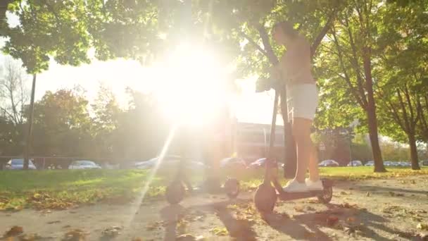 LOW ANGLE: Leende kvinnor njuta av en solig dag genom att rida e-skotrar genom parken — Stockvideo