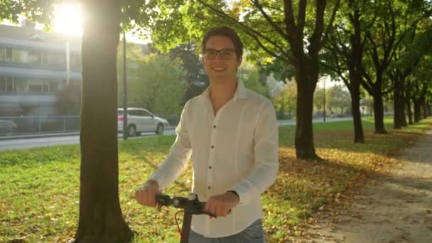 Unge man ler när han står över en e-scooter mitt i en allé. — Stockvideo