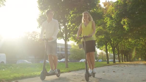 LENS FLARE: Leende kaukasiska par rider elektriska skotrar ner pulserande avenyn — Stockvideo