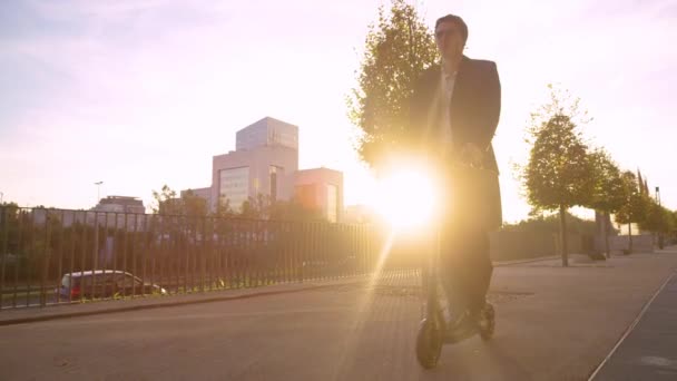 FLARE DE LA LENTE: Un hombre de negocios sonriente monta un e-scooter para trabajar en una mañana soleada . — Vídeo de stock