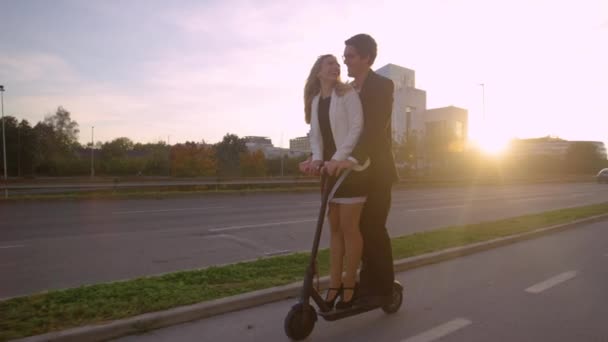 FLARE DE LA LENTE: Pareja joven en ropa formal montar un scooter eléctrico al atardecer . — Vídeos de Stock