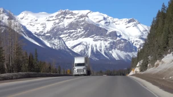 Trzy ciężarówki przewożą ładunek w dół autostrady prowadzącej w kierunku gór pokrytych śniegiem. — Wideo stockowe