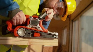 Sakallı inşaatçı inşaat alanında ahşap bir kirişi zımparalıyor.