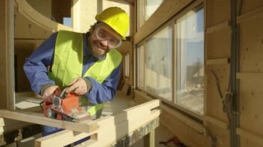 Gülümseyen inşaatçı prefabrik bir evde çalışıyor ahşap bir kiriş parlıyor..