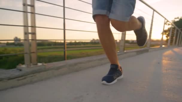 LOW ANGLE: Unrecognizable young man jogs across asphalt bridge at golden sunset. — Stock Video