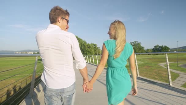 Happy young sweethearts holding hands cross a bridge on sunny day. — Stock video