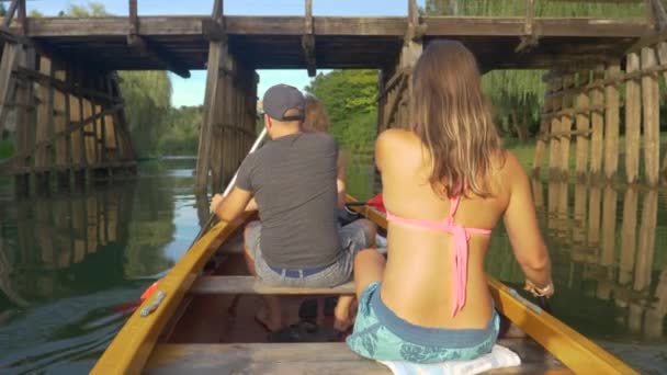 CERRAR: Turistas activos machos y hembras reman una canoa bajo un puente de madera. — Vídeo de stock