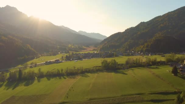 AERIAL: Złote letnie promienie słońca świecą na małej wiosce pod skalistą górą — Wideo stockowe