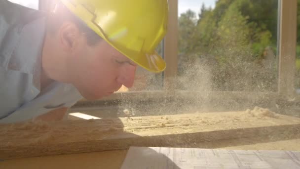 FERMER : Un jeune constructeur souffle une couche de sciure de bois sur une planche couchée sur un établi. — Video