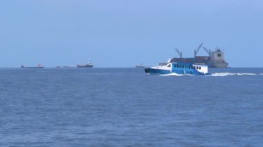 Mavi ve beyaz su taksisi yakınlardaki bir limandan gönderilen kargo gemilerinin yanından geçiyor.