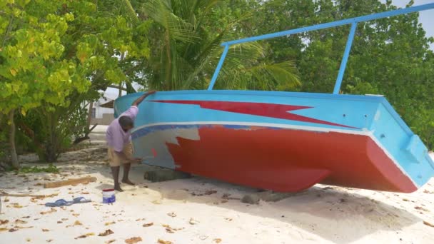 SLOW MOTION : Un pêcheur local peint son petit bateau en bois sur le rivage sablonneux. — Video