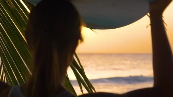 CERRAR: Mujer lleva una tabla de surf en la cabeza mientras camina a la playa al atardecer — Vídeo de stock