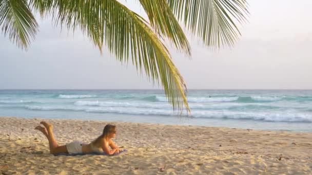 Młoda kaukaska kobieta leży na plaży i obserwuje surferów łapiących fale. — Wideo stockowe