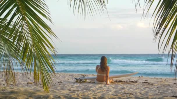 SLOW MOTION: Vrouw zit op zandstrand met een surfplank en kijkt naar de oceaan. — Stockvideo