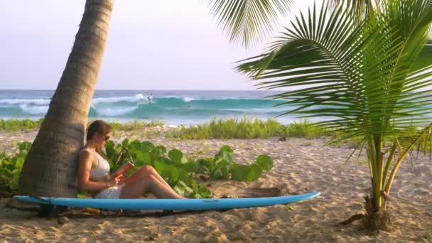 LOSE UP: Vrouw ontspant op het strand in de buurt van een populaire surfplek en leest een boek. — Stockvideo