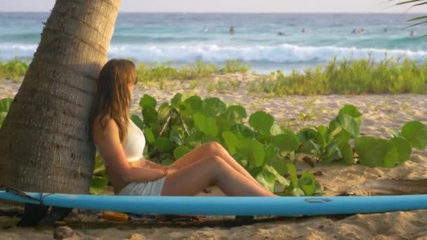 CLOSE UP Femme regarde les surfeurs monter les vagues tout en étant assis et appuyé sur le palmier — Video