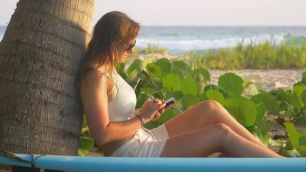 CLOSE UP: fiatal nő ül a strandon, közel egy szörf helyszínen, és SMS-t a barátainak. — Stock videók