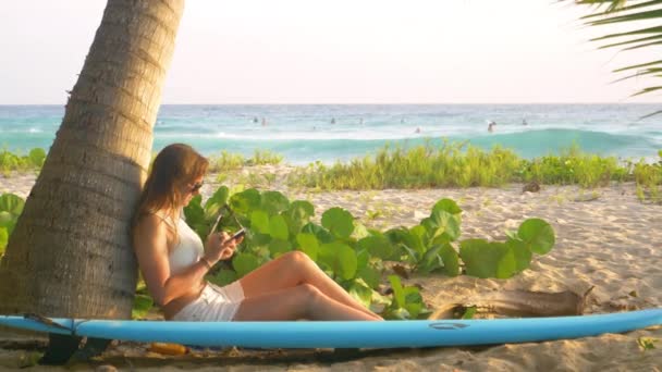 CLOSE UP: Молодий мандрівник спирається на пальму, сидячи на пляжі та смс — стокове відео