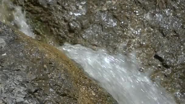 CLOSE UP: Скляний водоспад миє темно-коричневі та чорні камені в сонячний день — стокове відео