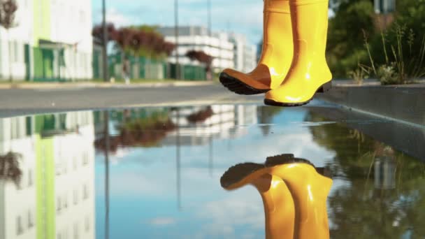 CLOSE UP: Carefree young woman in yellow rain boots jumps into the big puddle. — Stock Video