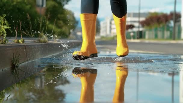 ÁNGULO BAJO: Mujer feliz con botas de lluvia amarillas corre a través de un charco vidrioso. — Vídeo de stock