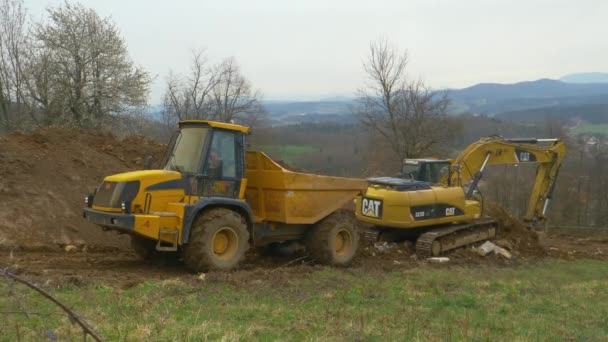 애벌레 굴착기가 노란 트럭 뒤에 있는 흙이 가득 담긴 양동이를 내리고 있다. — 비디오