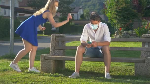 CLOSE UP: Girl wearing a facemask sprays man sitting on the bench and texting. — Stock Video
