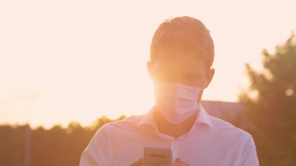 CLOSE UP: Young man wearing a medical facemask texts on a sunny summer evening. — Stock Video