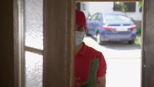 PORTRAIT: Kurier noszący maskę do twarzy podnosi pudełko produktów po otwarciu drzwi. — Wideo stockowe