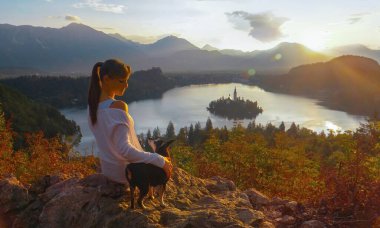 Neşeli kız köpeğiyle bir kayanın üzerinde oturuyor ve günbatımını seyrediyor..