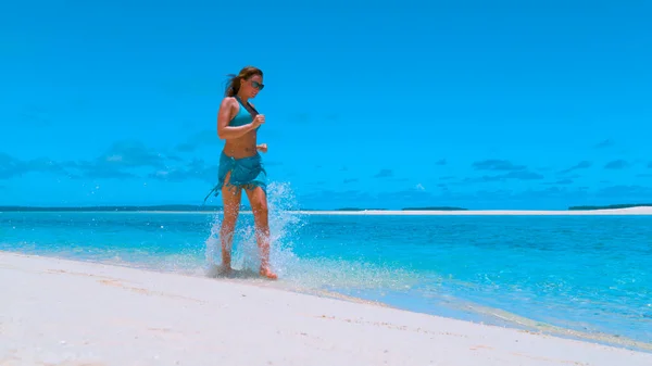 ANGOLO BASSO: Giovane donna sorridente in bikini che corre nell'acqua poco profonda dell'oceano. — Foto Stock