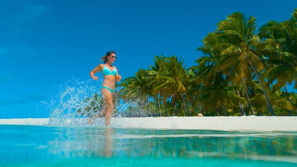 LOW Kąt: Uśmiechnięta dziewczyna rozbryzgująca szklaną wodę podczas biegania po plaży. — Zdjęcie stockowe