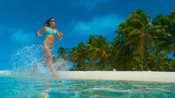 LOW Kąt: Szczęśliwa kobieta jogging w bikini i pluskania szklaną wodą oceanu. — Zdjęcie stockowe