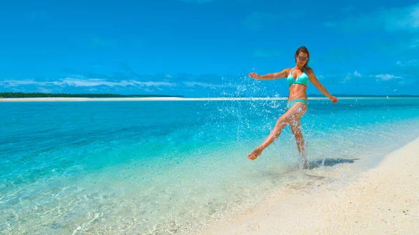 ÁNGULO BAJO: Mujer joven feliz en bikini pateando y salpicando agua cristalina del océano. — Foto de Stock