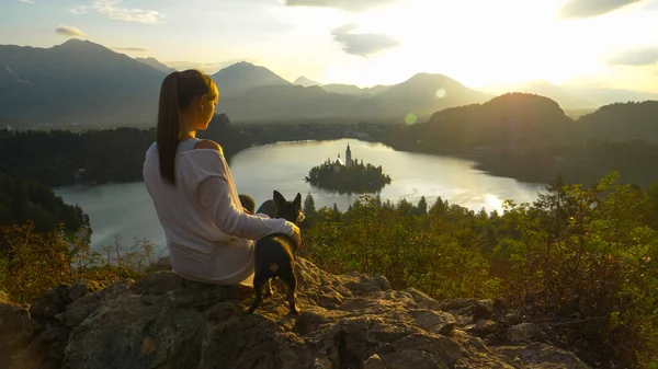 ΚΛΕΙΣΙΜΟ: Κορίτσι κάθεται στο βράχο και βλέποντας το ηλιοβασίλεμα πάνω Bled με το σκυλί. — Φωτογραφία Αρχείου