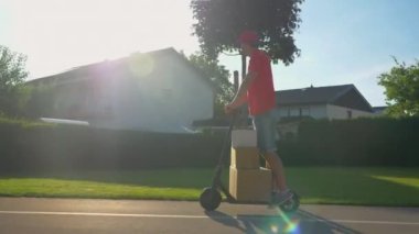Genç kurye güneşli bir günde e-scooterıyla paket teslim ediyor..