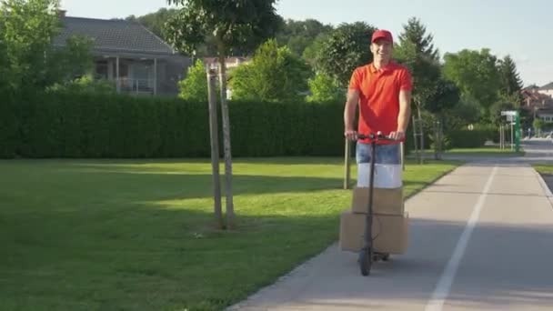El mensajero sonriente monta un scooter eléctrico mientras entrega paquetes en los suburbios — Vídeos de Stock
