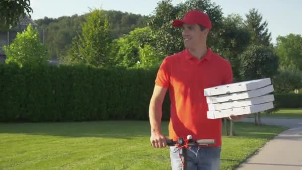 Kırmızı tişörtlü kurye e-scooterla pizza dağıtırken gülümsüyor. — Stok video