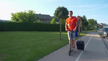 Mutlu çift e-scooter kullanıyor ve bagajlarını arkalarından sürüklüyor..