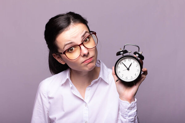 girl in a white shirt and glasses holds an alarm clock in her hand
