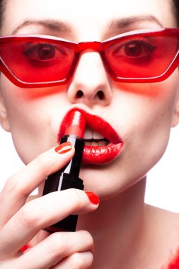 Kırmızı kazaklı ve kırmızı gözlüklü kız dudaklarını kırmızı rujla boyuyor.