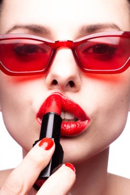 Kırmızı kazaklı ve kırmızı gözlüklü kız dudaklarını kırmızı rujla boyuyor.