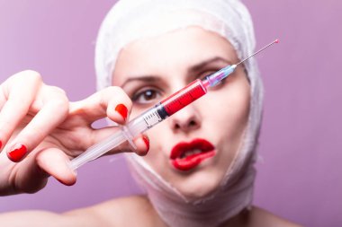 Güzel genç bir kız, dudakları kırmızı rujla boyanmış, kafası bandajlı, elinde kırmızı bir sıvıyla bir şırınganın