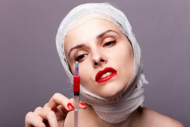 Güzel genç bir kız, dudakları kırmızı rujla boyanmış, kafası bandajlı, elinde kırmızı bir sıvıyla bir şırınganın