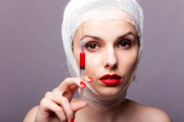 Güzel genç bir kız, dudakları kırmızı rujla boyanmış, kafası bandajlı, elinde kırmızı bir sıvıyla bir şırınganın